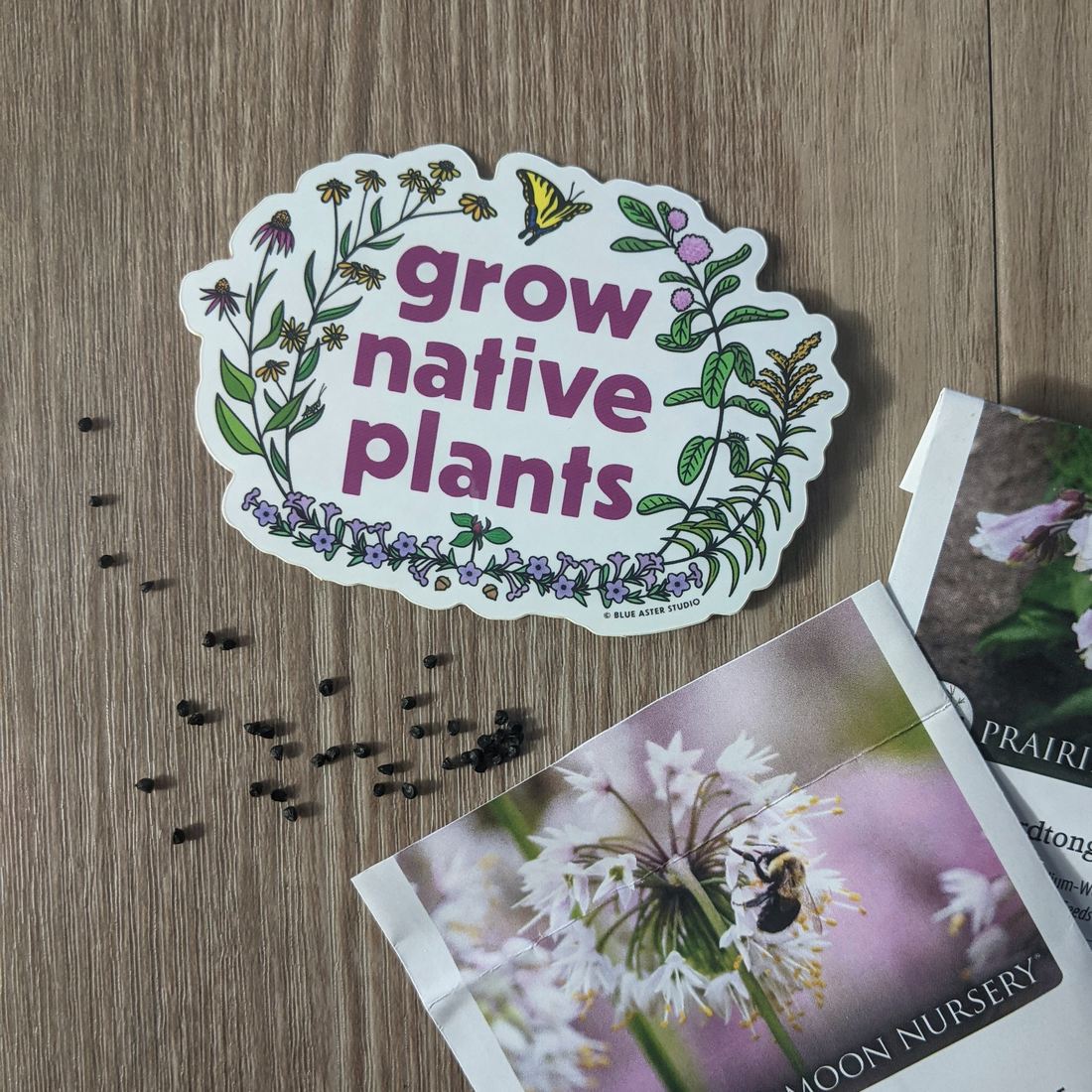A grow native plants sticker sitting among Prairie Moon Nursery seed packets. Some nodding onion seeds are scattered on the table top.