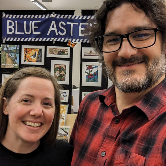 Selfie of Jennie and David in front of the Blue Aster Studio booth featuring many screen prints of wildlife