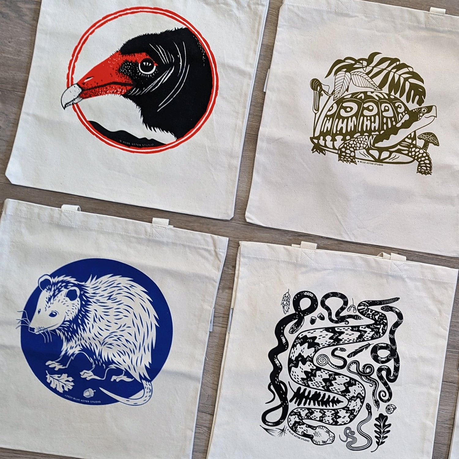 Blue Aster Studio tote bags with illustrations of wildlife including a turkey vulture, box turtle, opossum, and assorted snakes