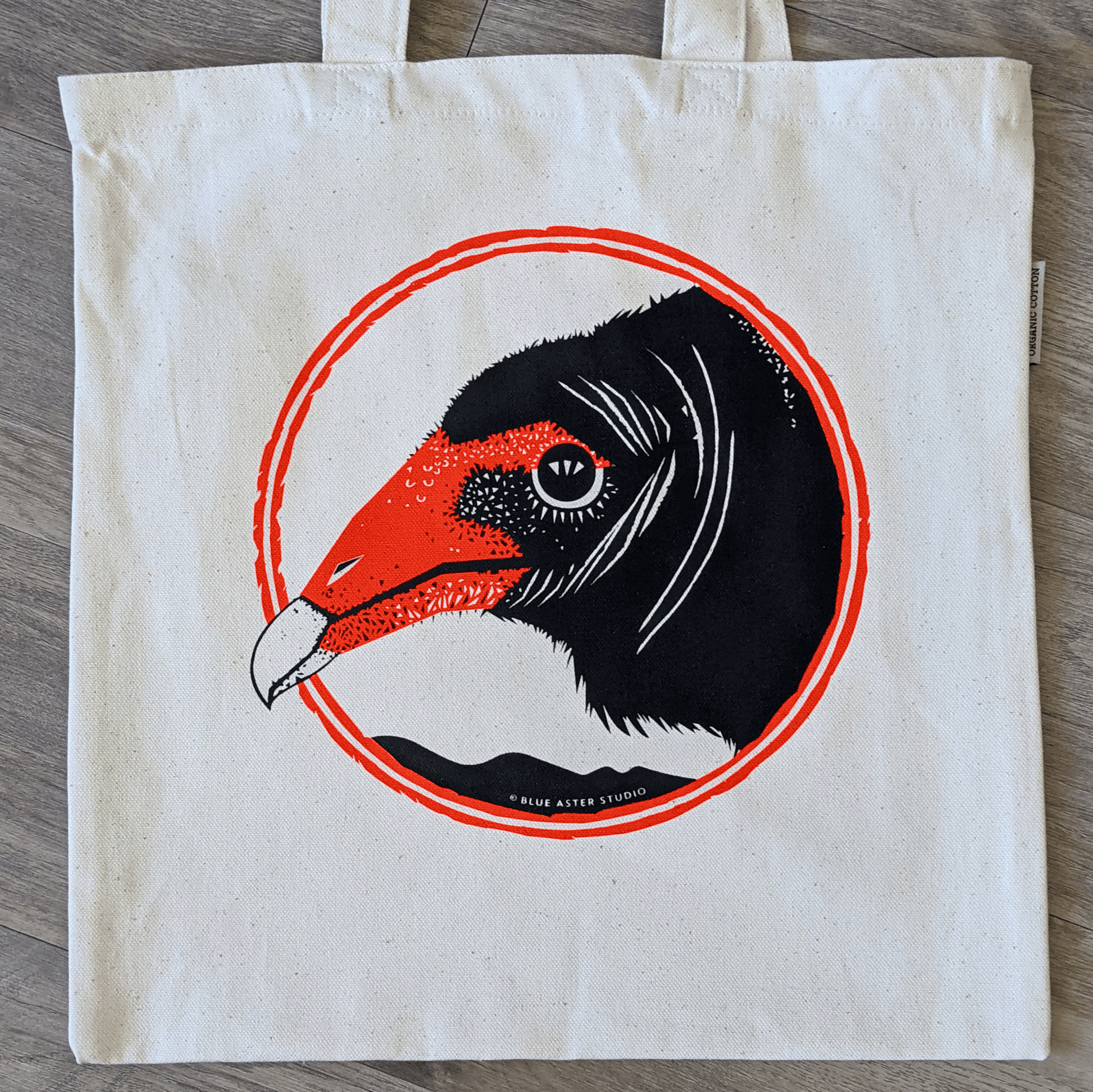 An organic cotton turkey vulture tote bag. The bag has a turkey vulture face screen printed on it in black and red.