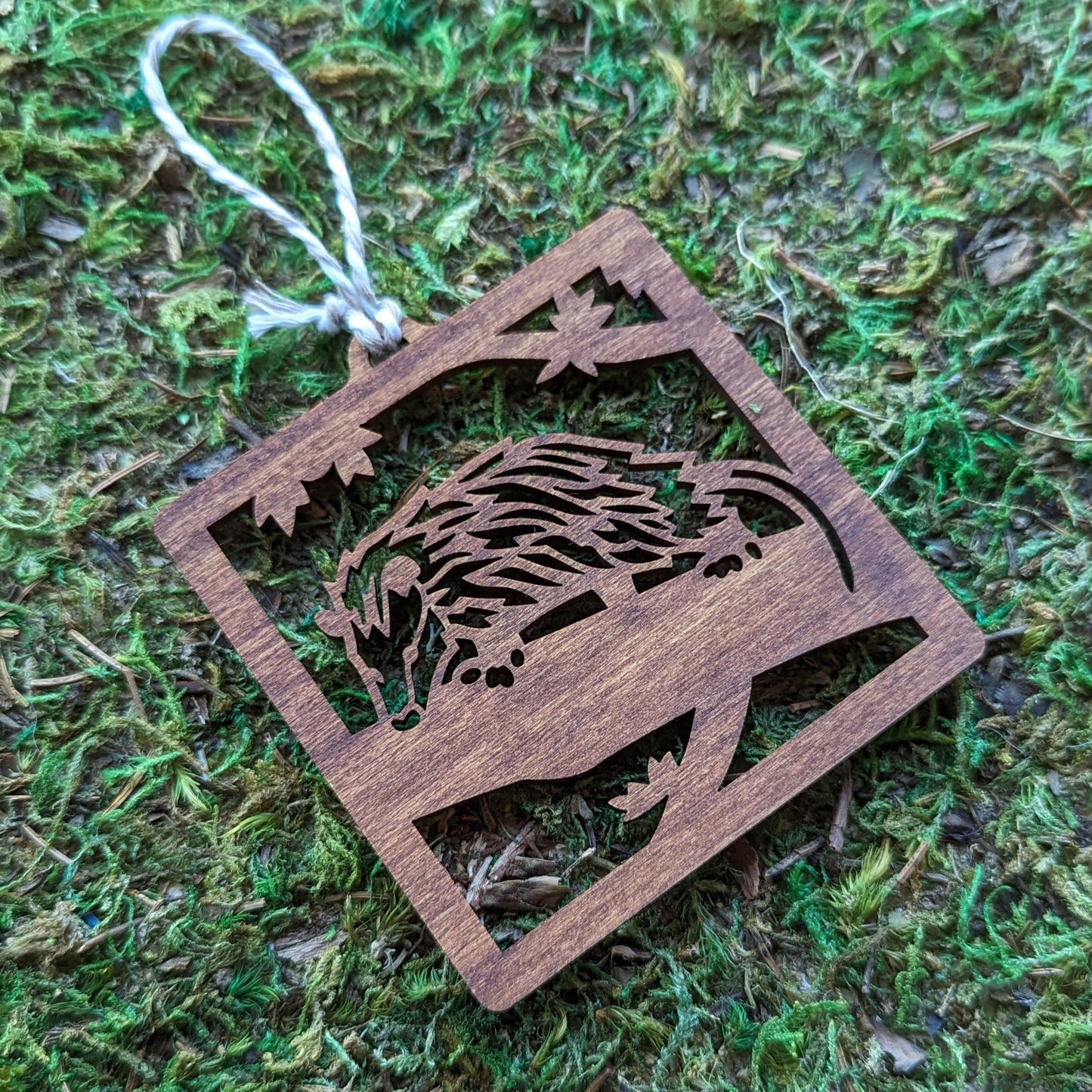 A wooden laser cut ornament of an opossum on a tree branch colored with brown wood dye on a mossy background.
