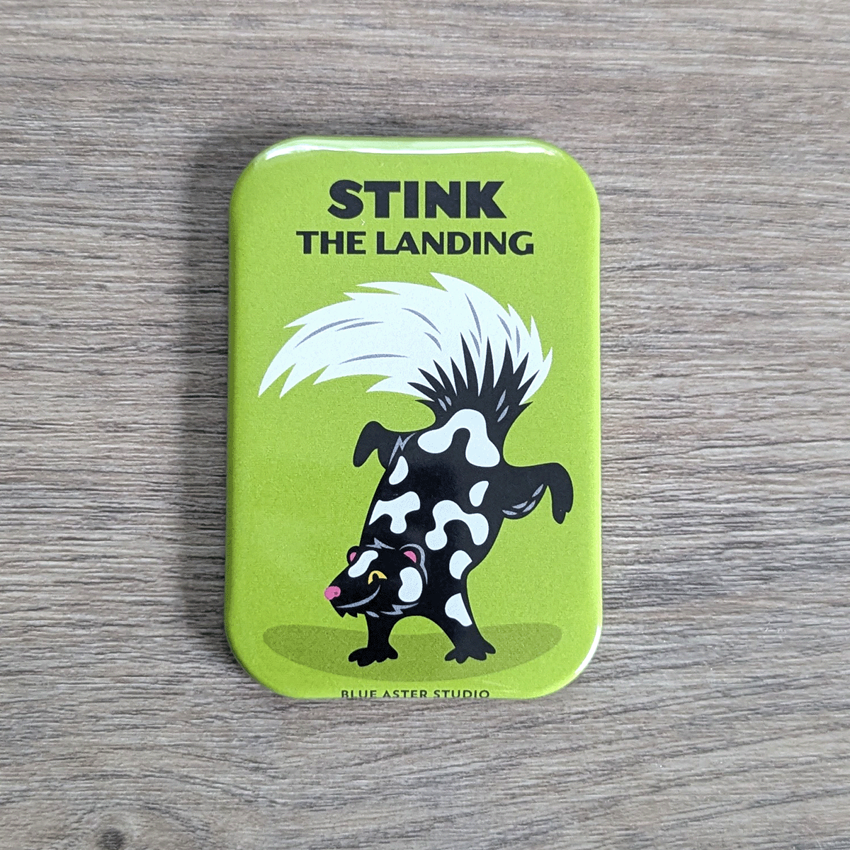 A rounded rectangle magnet featuring an illustration of a spotted skunk doing a hand stand under the text Stink the Landing.