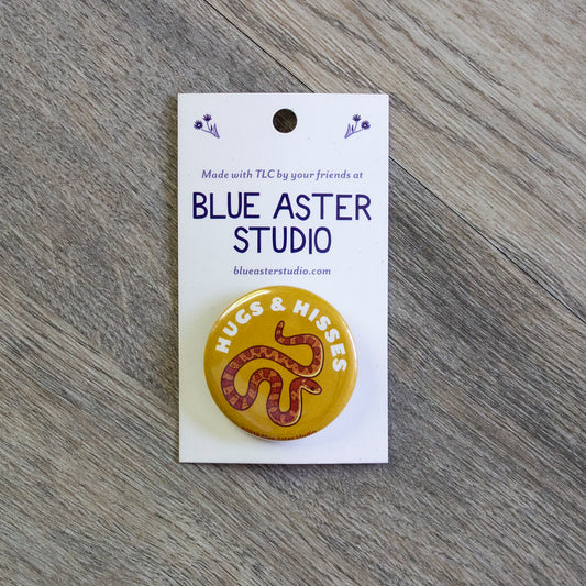 A 1.5 inch yellow pinback button with an illustration of a corn snake and the words Hugs And Hisses.