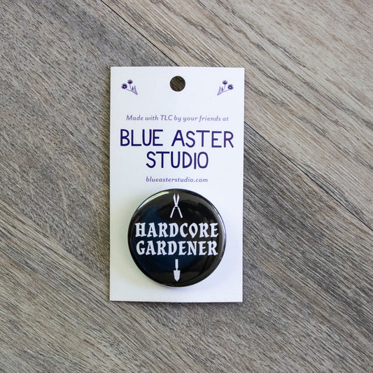 A 1.5 inch black pinback button with the words Hardcore Gardener in white. There are white silhouettes of gardening shears above the words and a gardening trowel below.