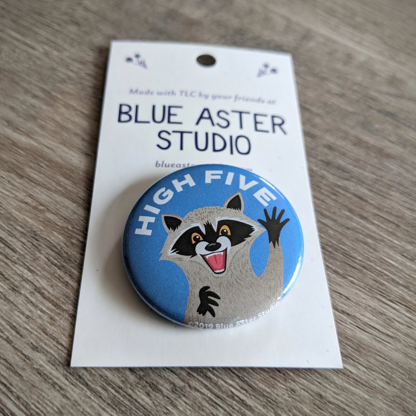 A close up of the 1.5 inch button with an illustration of a raccoon with its hand up with the words "High Five" above it.