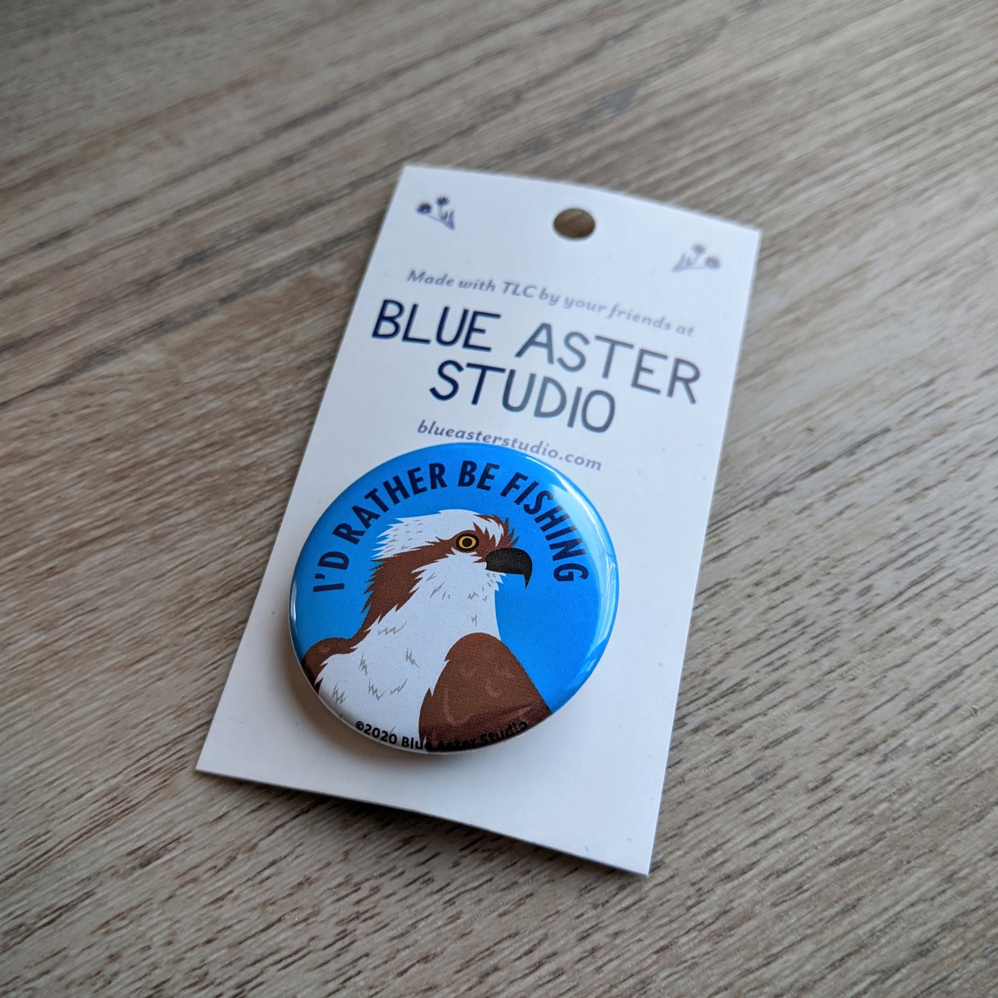 A 1.5 inch pinback button with an illustration of an osprey and the words "I'd Rather Be Fishing"