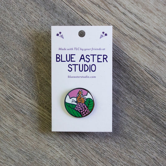 A circular enamel pin of a monarch butterfly perched on the pink flower of a common milkweed plant with white clouds and a pink sunset sky behind it.