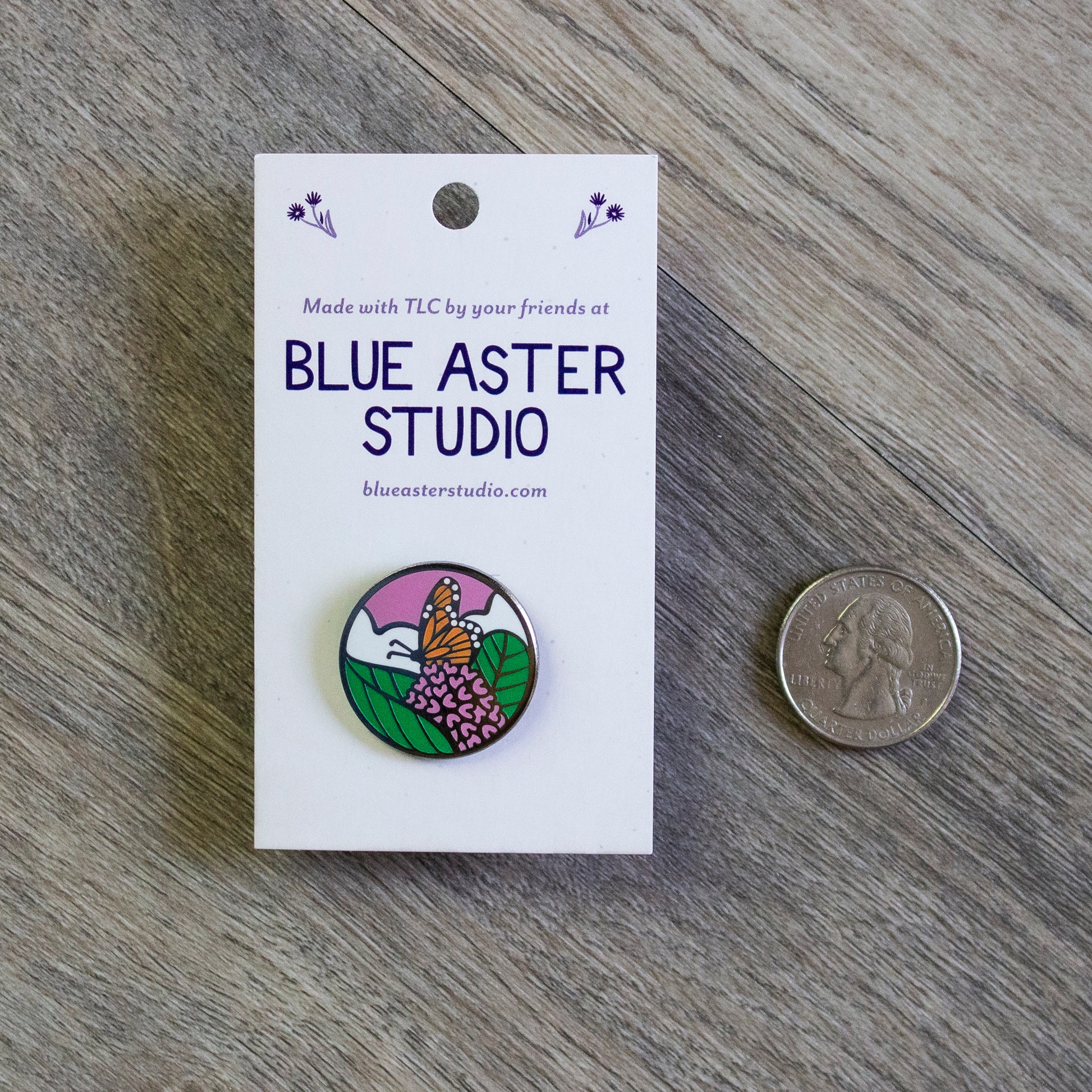 A circular enamel pin of a monarch butterfly perched on the pink flower of a common milkweed plant with white clouds and a pink sunset sky behind it. A US quarter is provided for scale.