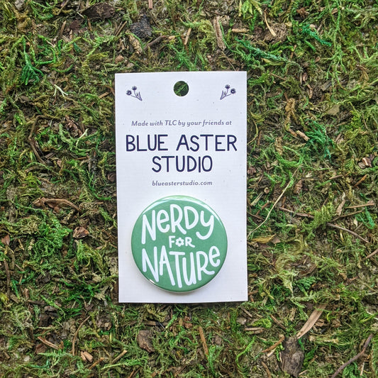 Hand holding a 1.5 inch pinback button on a card. The button has the words "Nerdy For Nature" in white on a teal background.