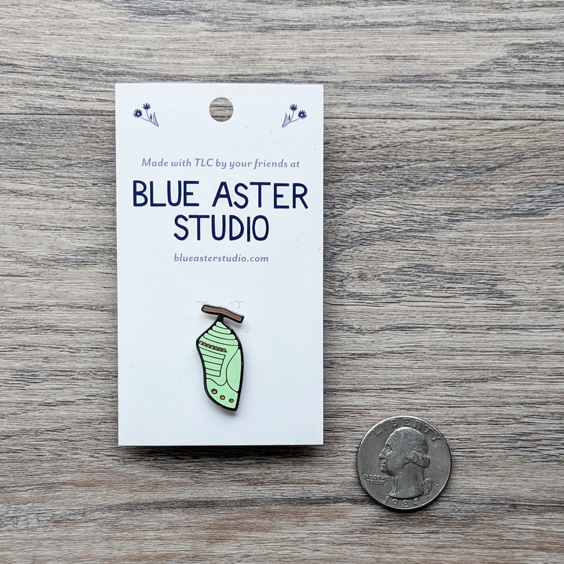 A monarch butterfly chrysalis enamel pin next to a USD quarter for scale.