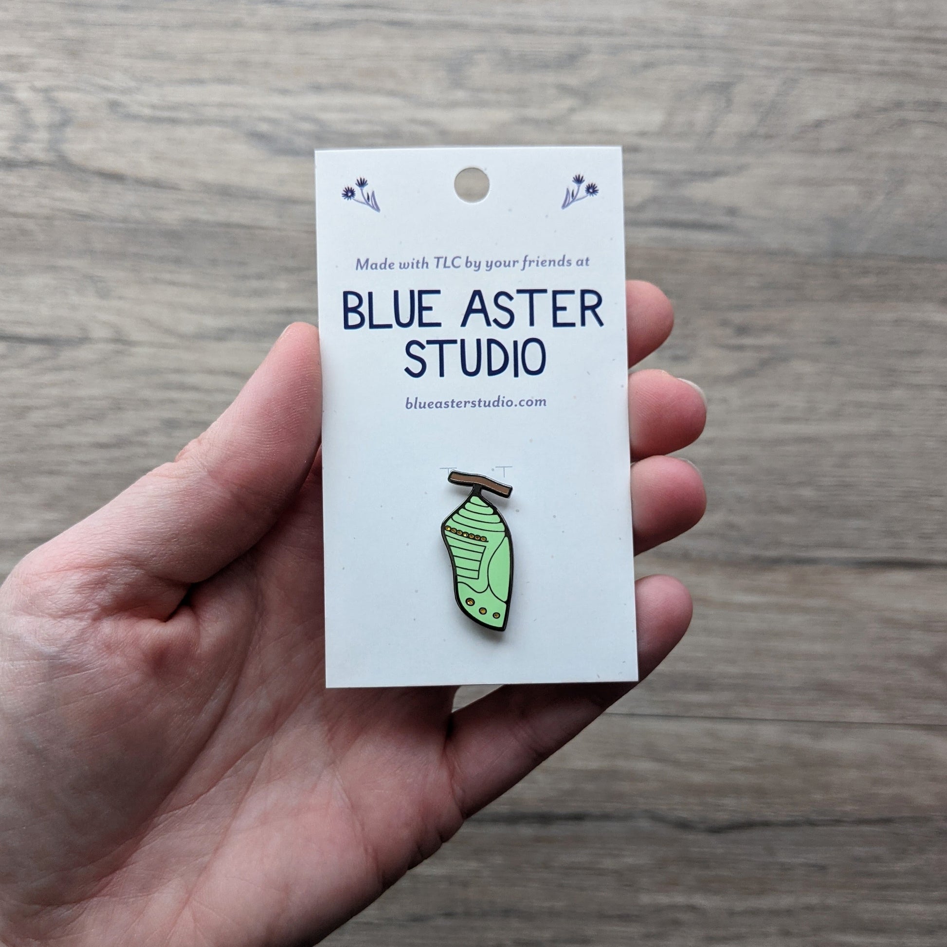 An enamel pin in the shape of a monarch butterfly chrysalis being held by a hand to show scale.