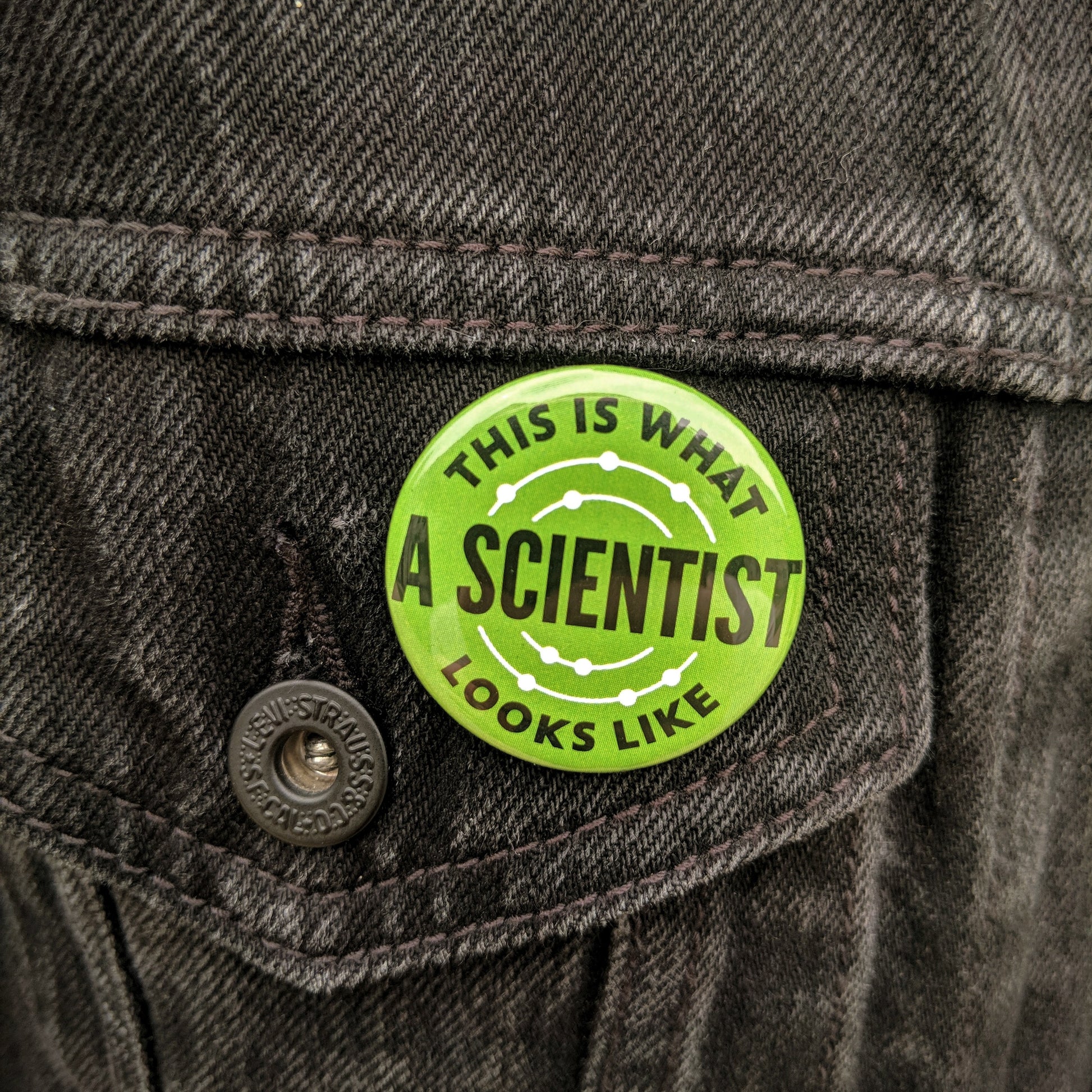 The green This Is What A Scientist Looks Like button pinned on a black jean jacket.
