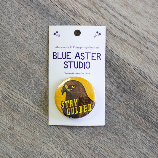 A 1.5 inch pinback button with an illustration of a golden eagle with a sunburst of yellow and orange behind it and the words Stay Golden.