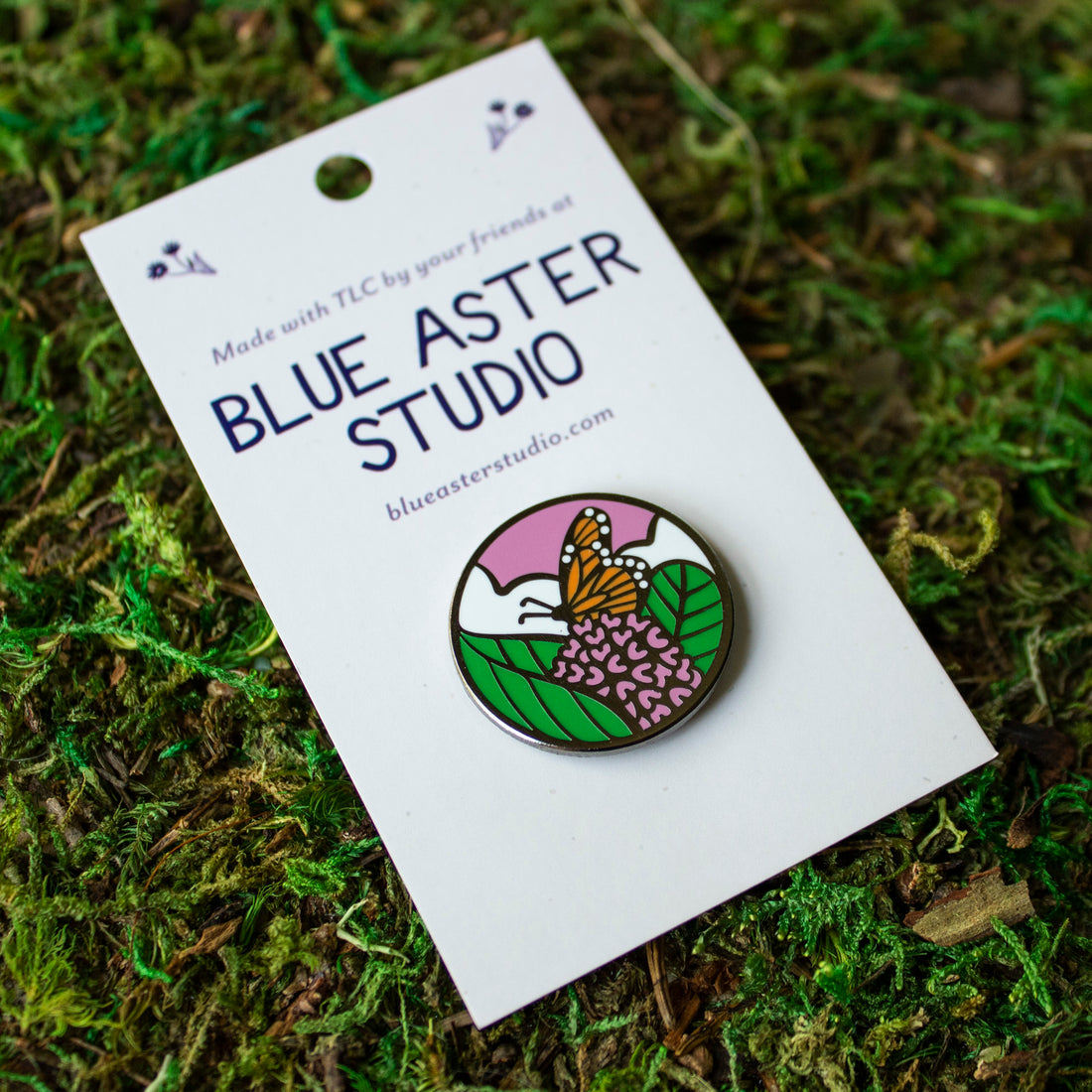 An enamel pin of a monarch butterfly nectaring on the flowers of the common milkweed plant.