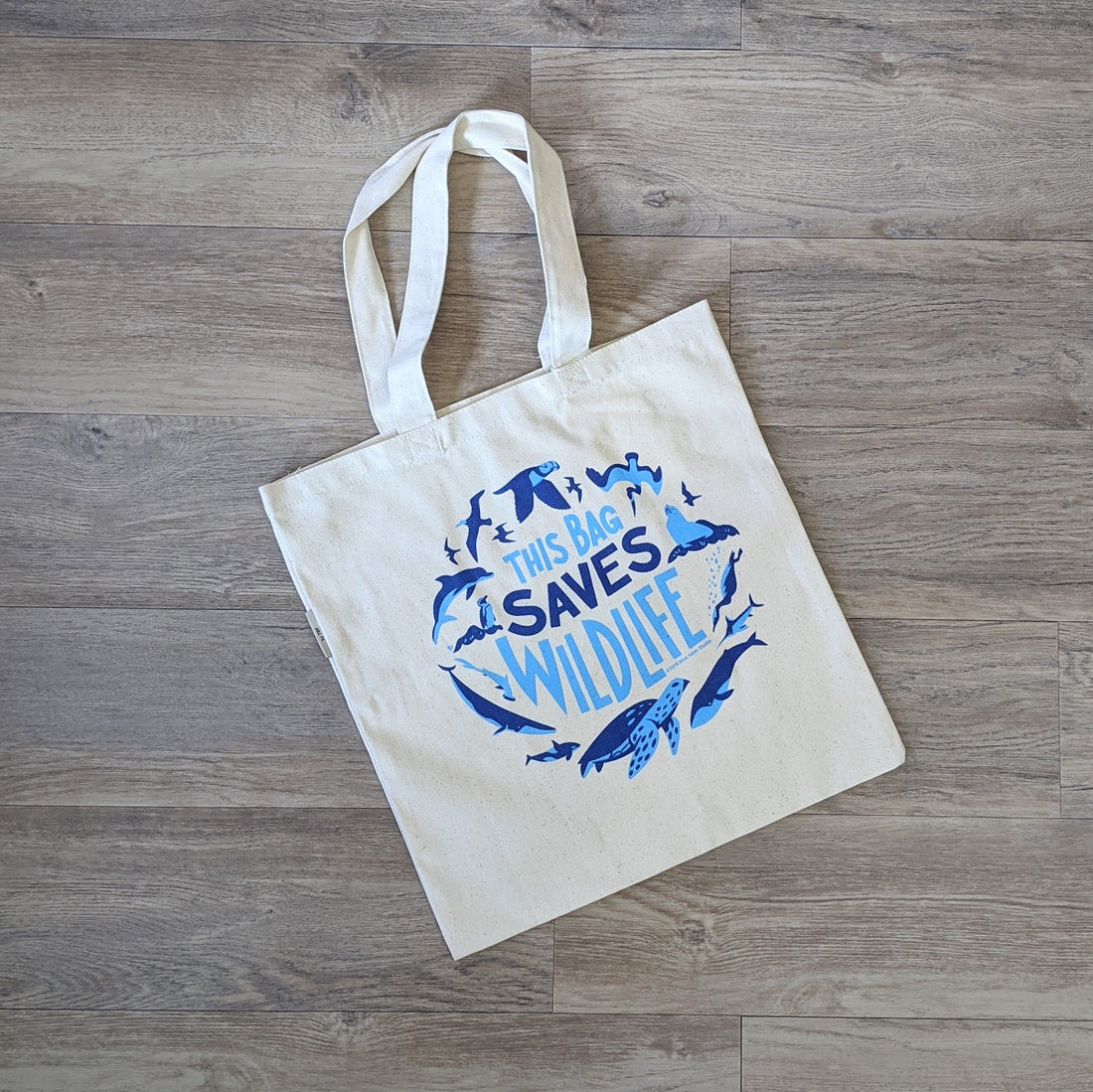 An organic cotton tote bag with the words "This Bag Saves Wildlife" with illustrations of ocean wildlife surrounding it screen printed in two shades of blue