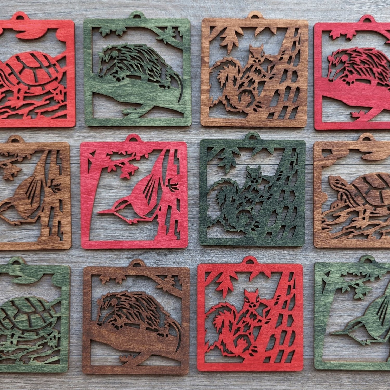 A grid layout of wooden wildlife ornaments in green, brown, and red. Animals include a nuthatch, squirrel, opossum, and painted turtle.