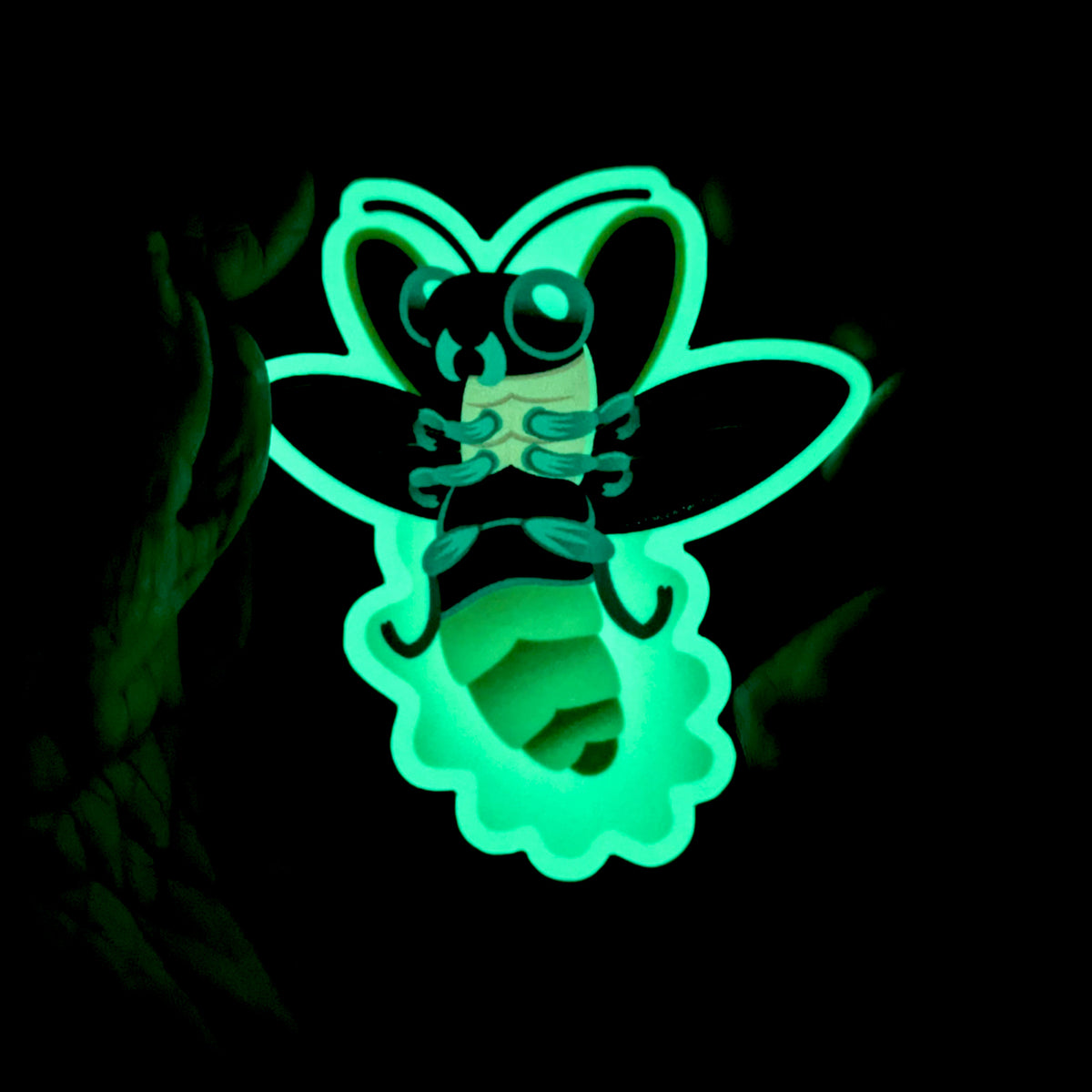 An illustrated sticker of a firefly in flights with a glowing abdomen, held in hand in the dark to show the brilliant green glow of the sticker