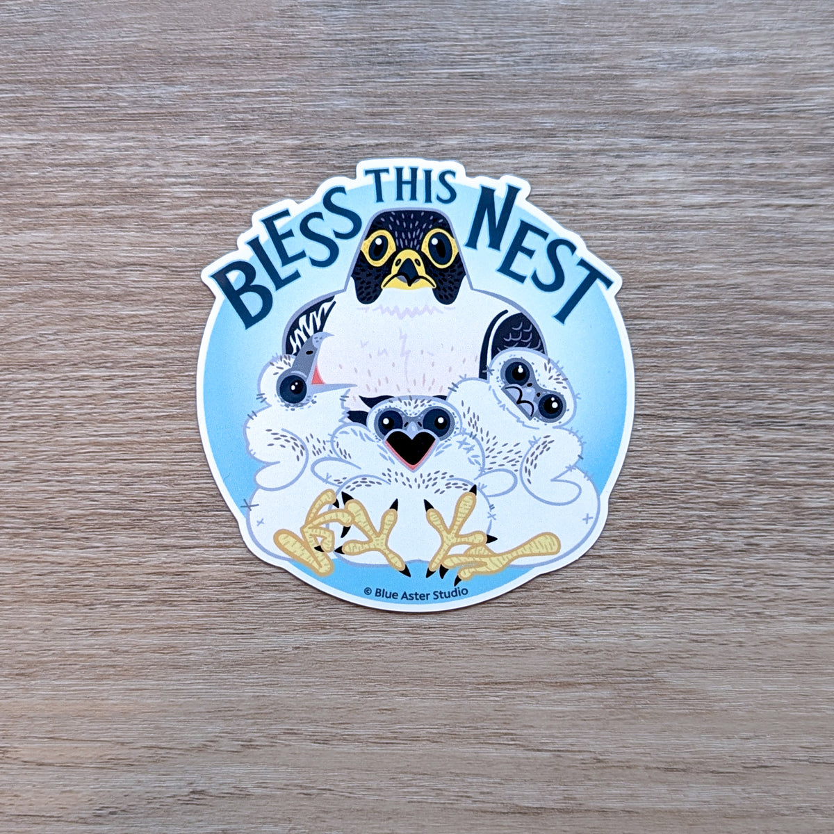 A round sticker depicting an illustrated parent peregrine falcon with three chicks and the text "Bless This Nest"