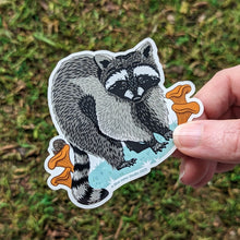 Load image into Gallery viewer, The raccoon vinyl sticker being held in a hand.