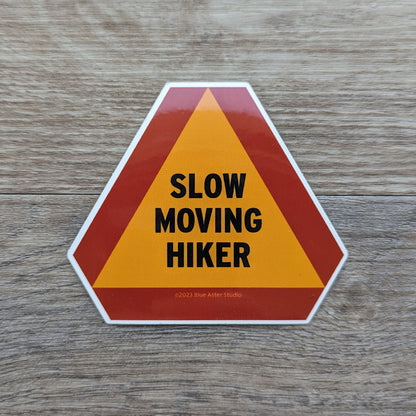 A vinyl sticker with the words "Slow Moving Hiker" in an orange triangle with a reddish orange border.