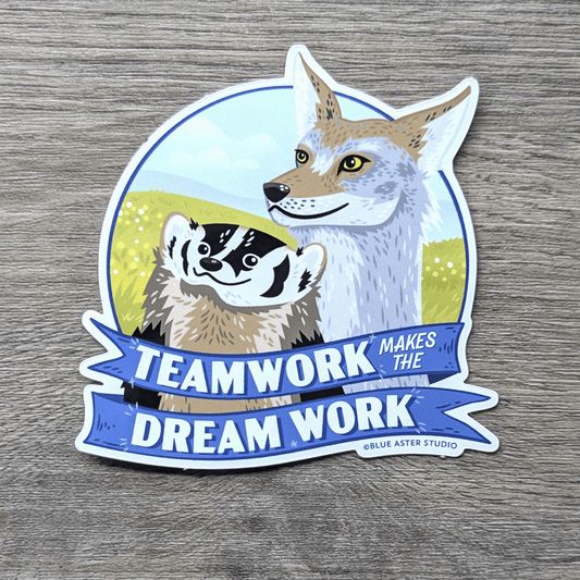 A sticker featuring an illustration of an American badger and a coyote smiling and looking to the horizon in front of a grassland. A blue ribbon below them reads Teamwork Makes the Dream Work.