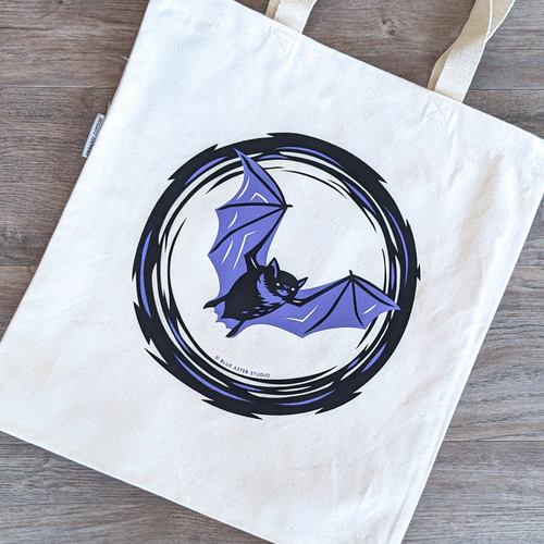 A sturdy organic cotton canvas tote bag with a flying bat design screen printed on it in purple and black inks.