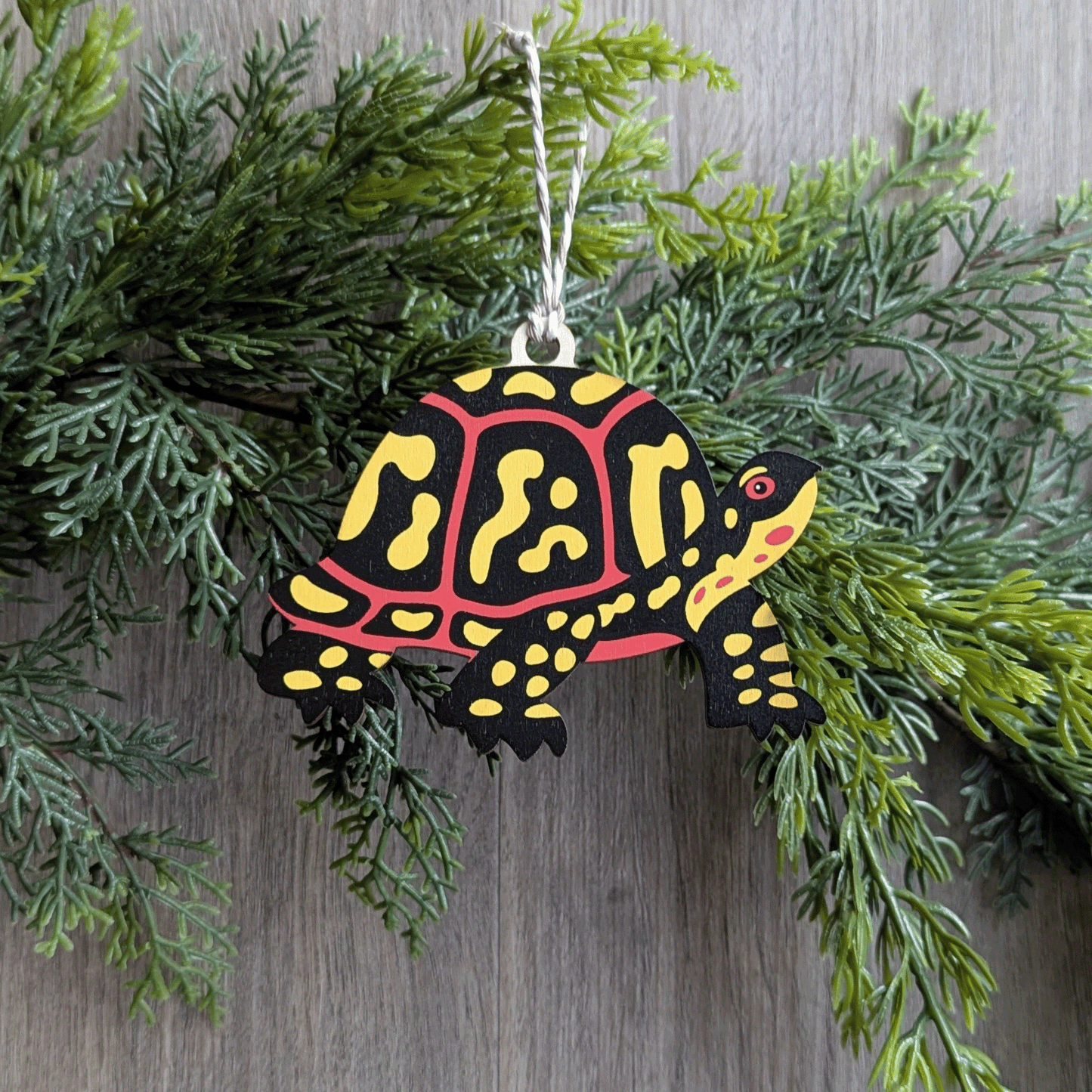 A wooden screen printed ornament of a box turtle hanging on a hemlock garland.