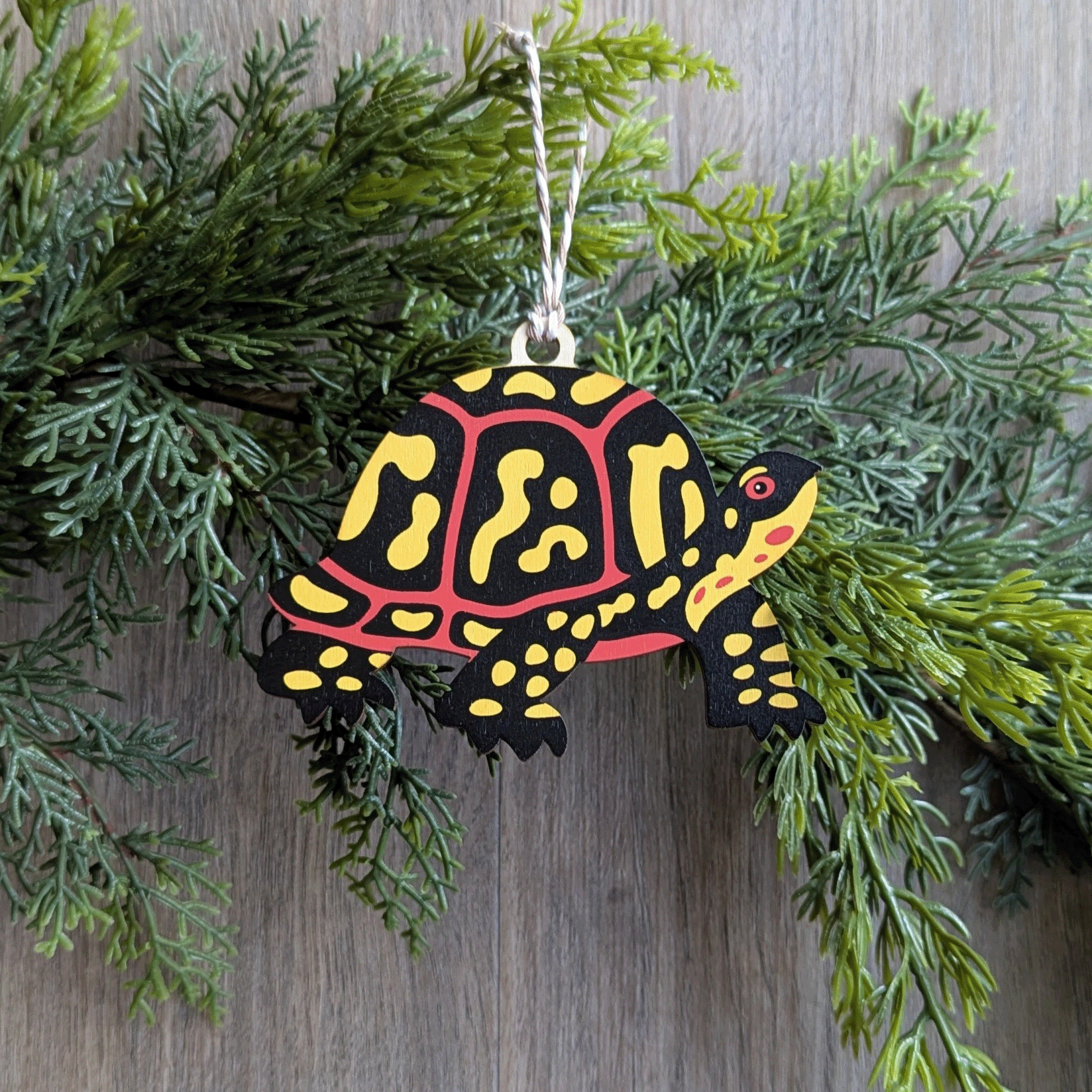 A wooden screen printed ornament of a box turtle hanging on a hemlock garland.