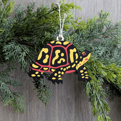 A wooden screen printed ornament of a box turtle hanging on a hemlock garland.