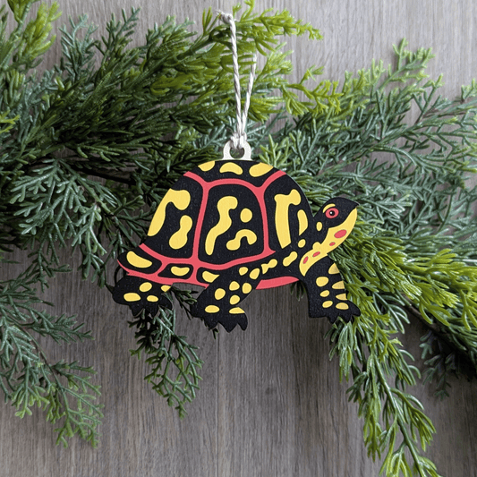 A wooden screen printed ornament of a box turtle hanging on a hemlock garland.