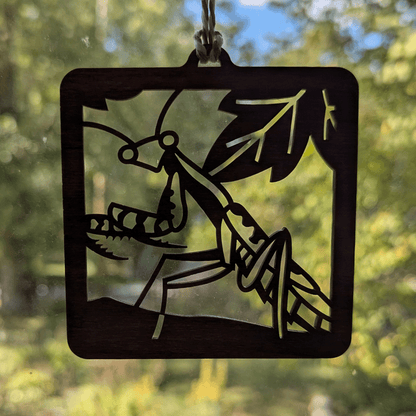 A square wooden ornament of a praying mantis in front of a window.
