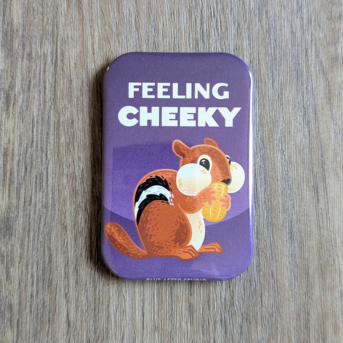 A two inch by three inch rectangular magnet featuring an illustrated chipmunk holding a peanut, set against a purple background. The text above the chipmunk reads "Feeling Cheeky."