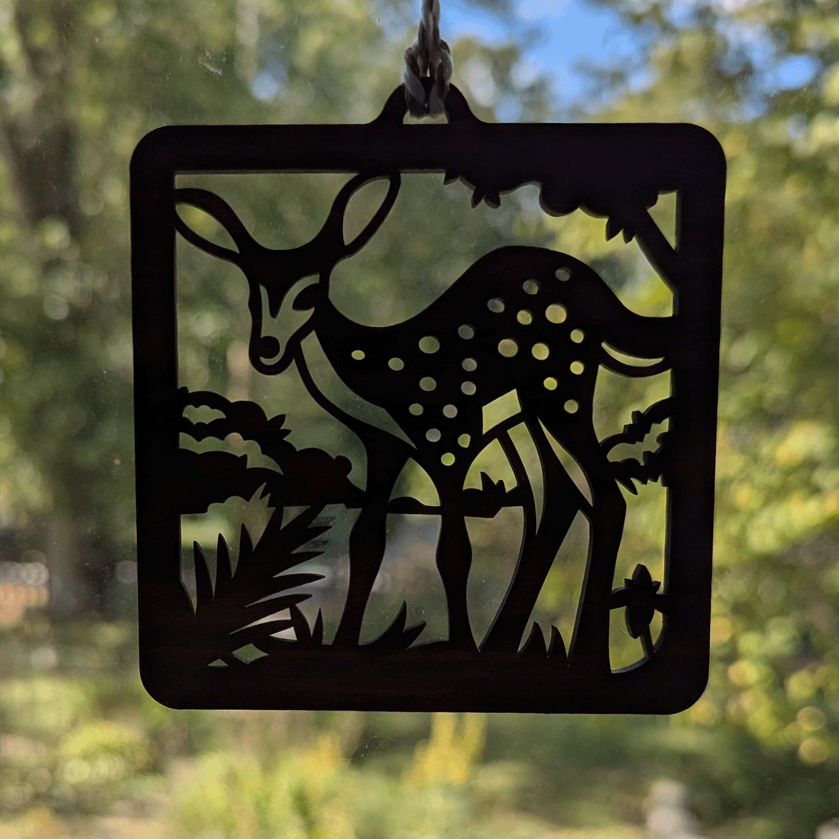 A square wooden ornament of a spotted fawn hung in a window.