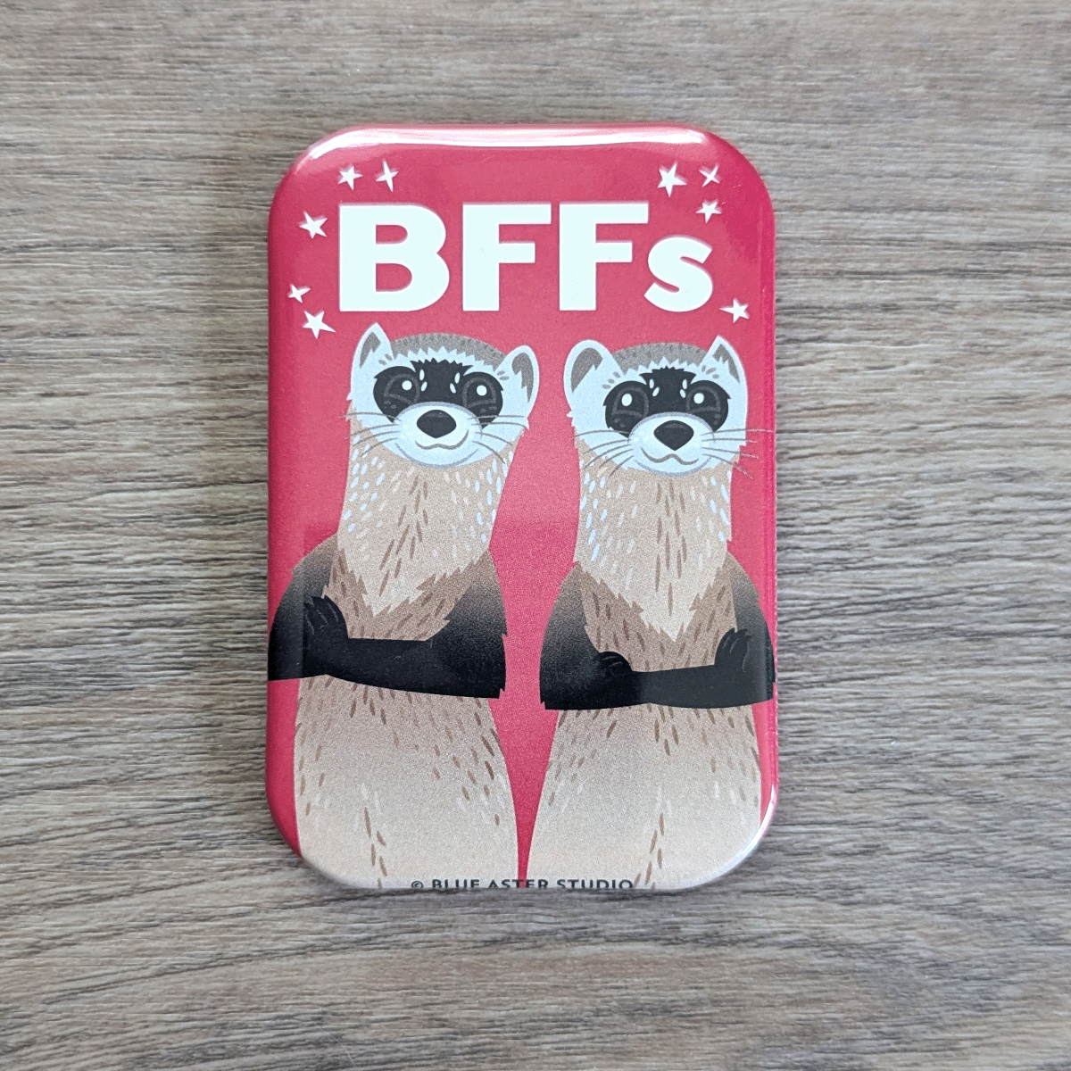A rounded rectangle magnet featuring an illustration of two black-footed ferrets standing back to back and smiling with the text BFFs above them.
