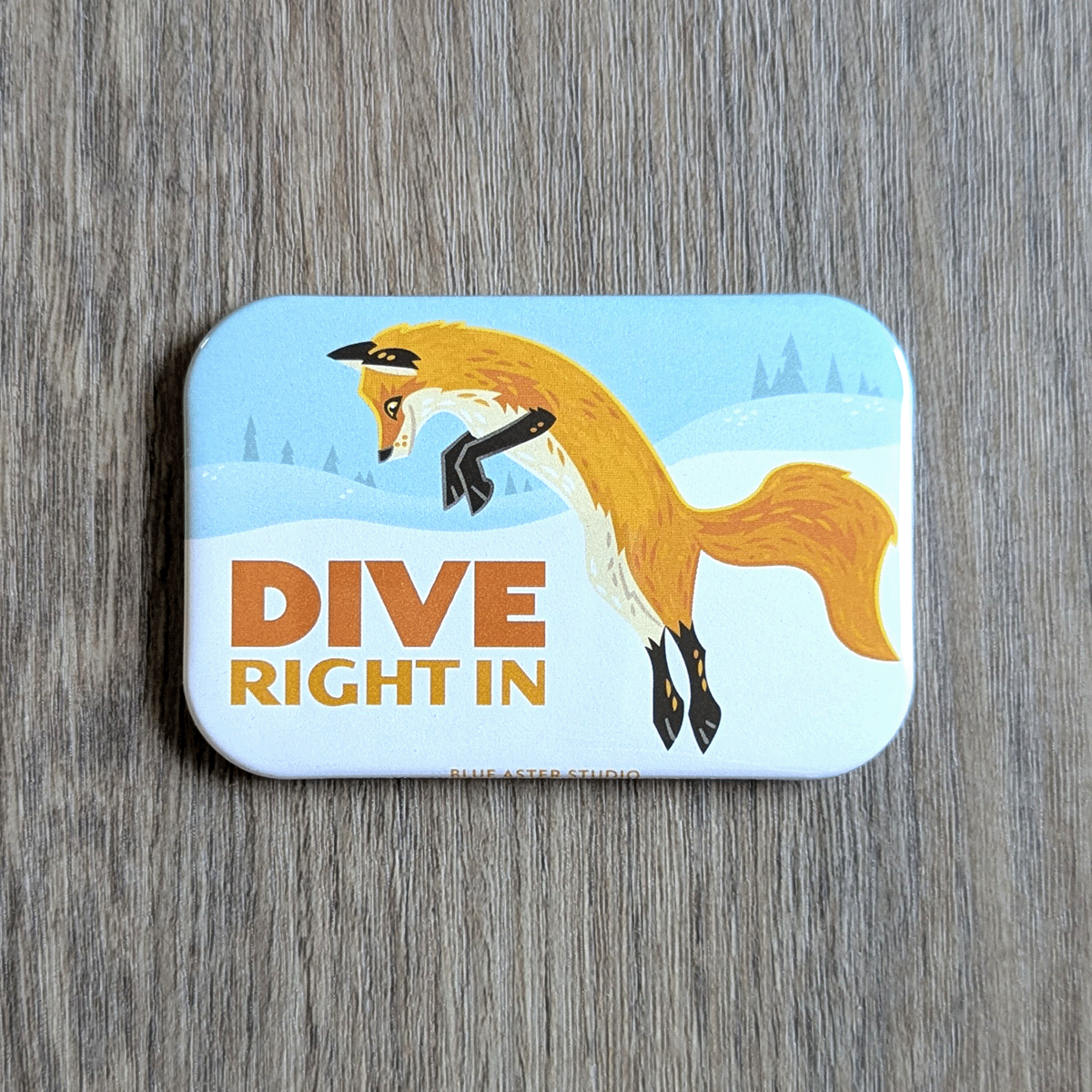 A rectangular magnet with an illustration of a red fox diving into the snow with the text dive right in