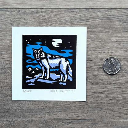 A square screen printed artwork of a gray wolf standing on a flat rock with mountains in the distance. A full moon shines above. A USD quarter is included to show the size of the four inch square print.