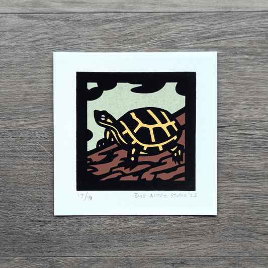 Screen printed art of a yellow and black turtle on a log with green water and lily pads around it.