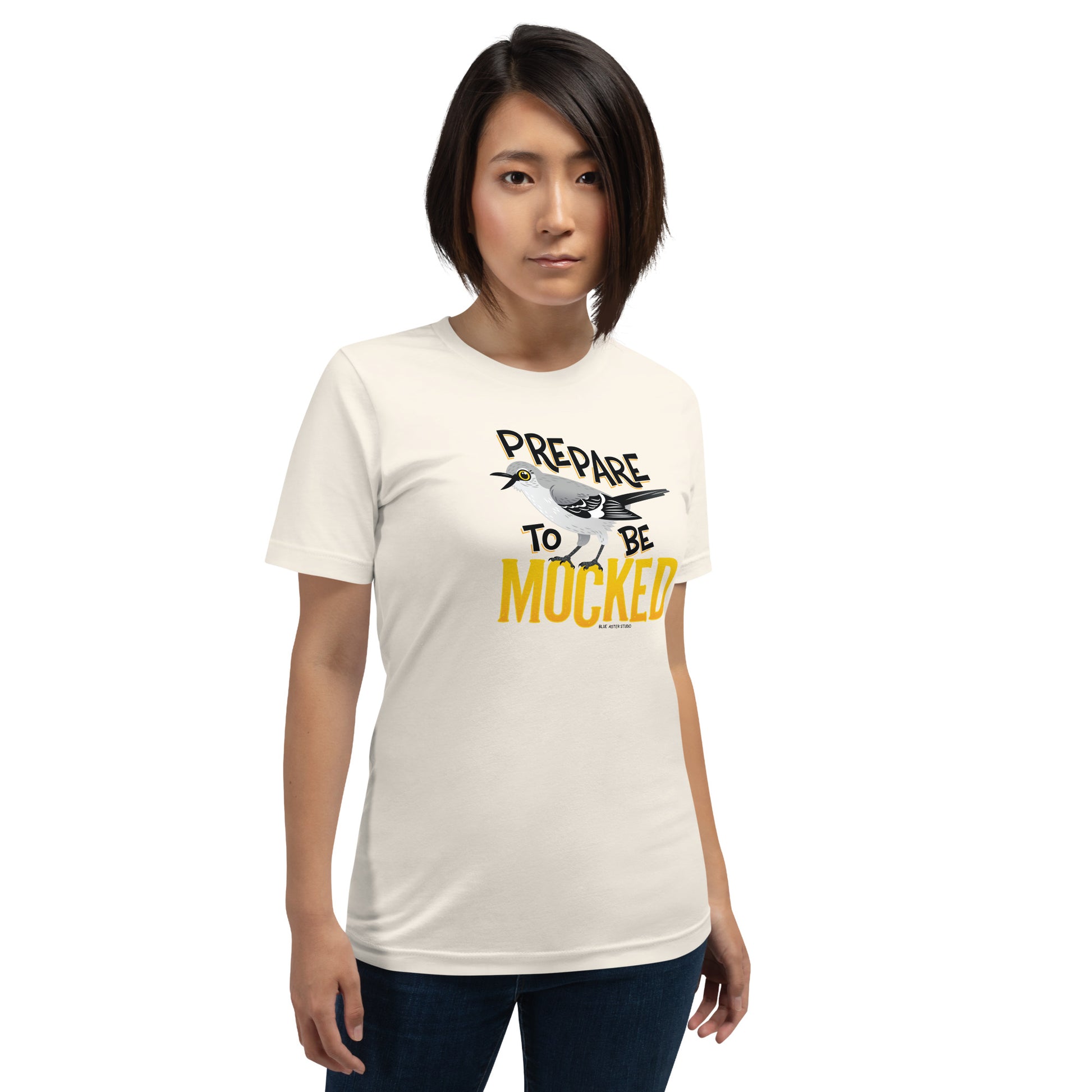 A creme tee shirt worn by a female model with the text "prepare to be mocked" with an illustrated mockingbird perched within it