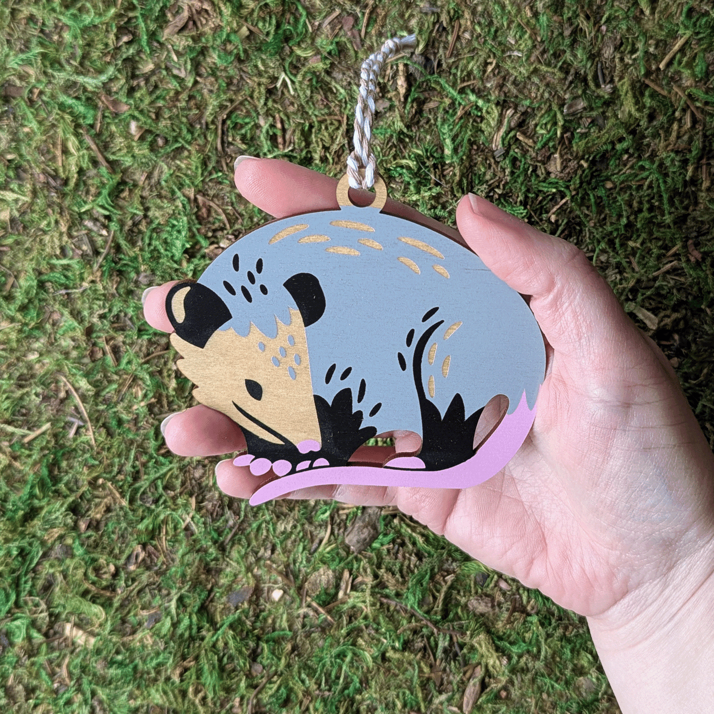 A screen printed wooden ornament of a Virginia opossum held in a hand, it is about the size of the palm.