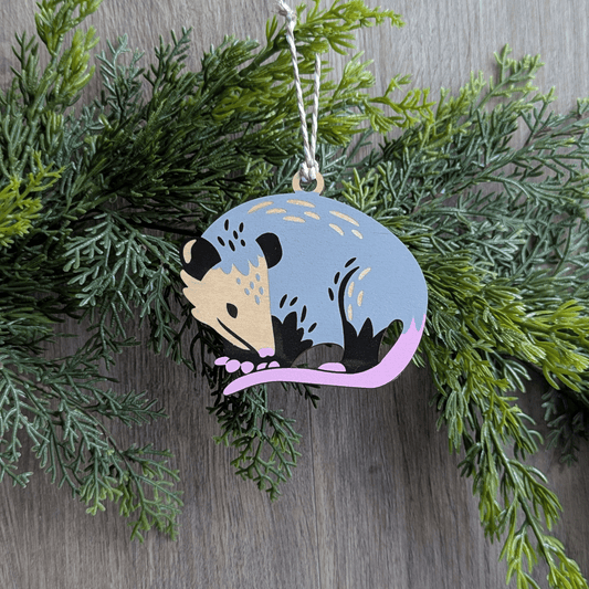 A screen printed wooden ornament of a Virginia opossum hanging on a green hemlock garland