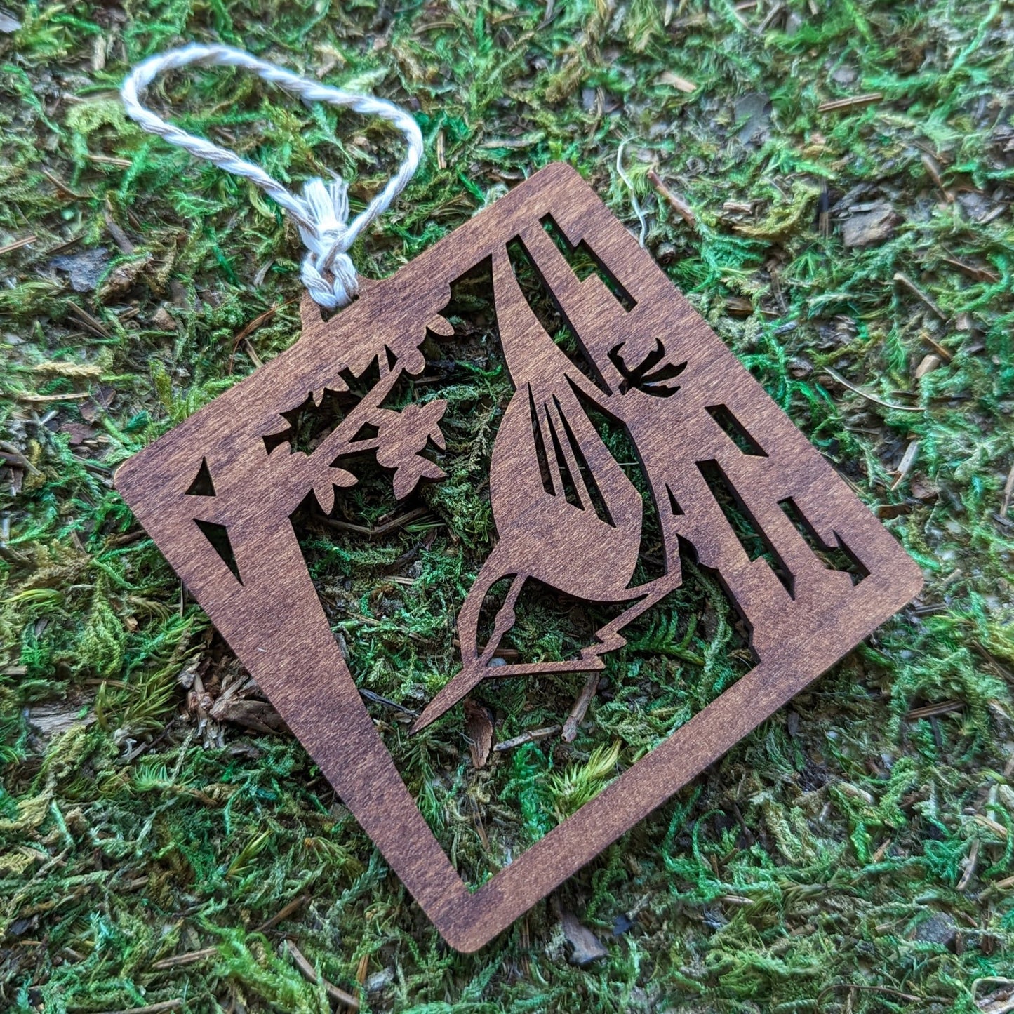 A wooden laser cut ornament of an opossum on a tree branch colored with brown wood dye on a mossy background.