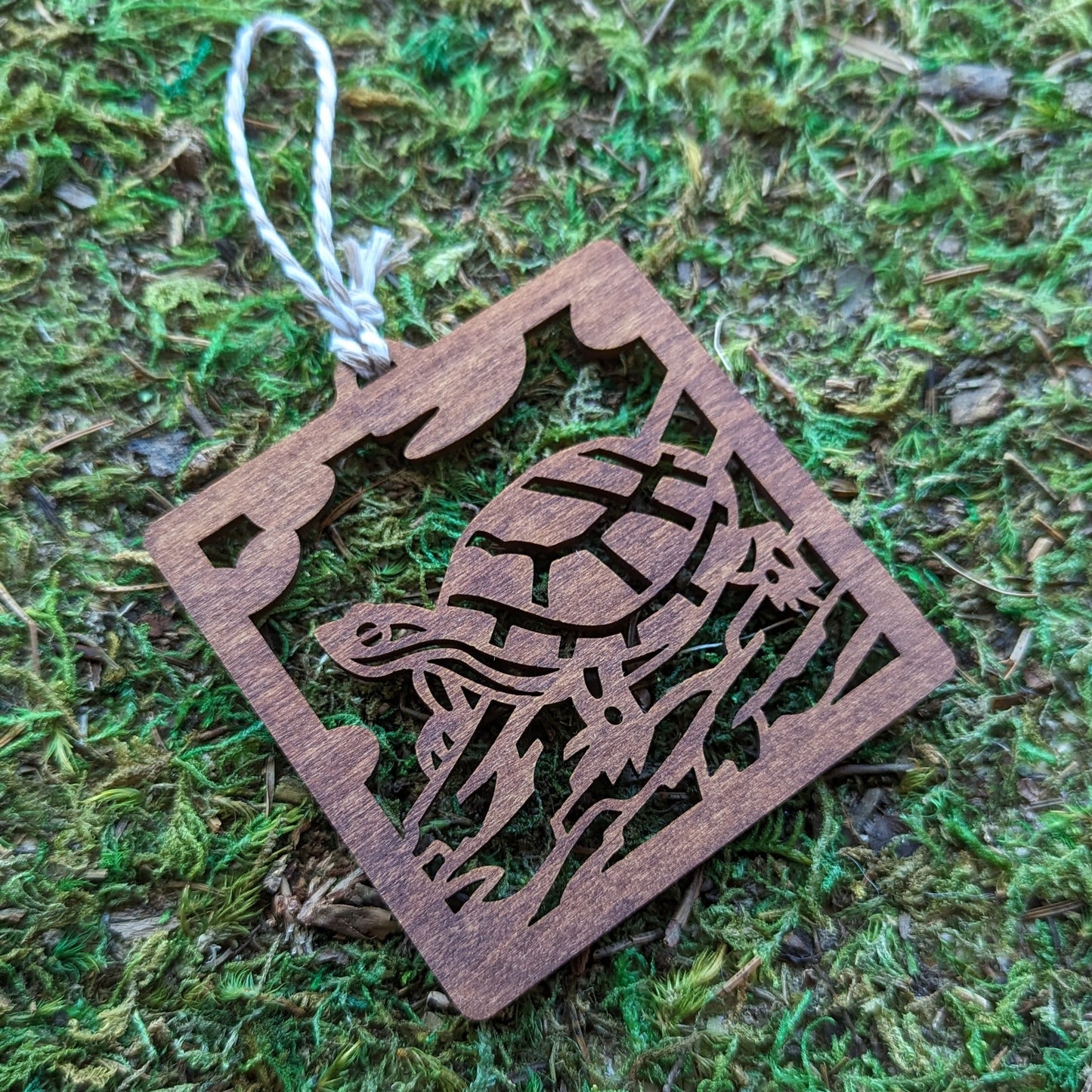 A wooden laser cut ornament of a turtle sitting on a log colored with brown wood dye on a mossy background.