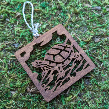 A wooden laser cut ornament of a turtle sitting on a log colored with brown wood dye on a mossy background.