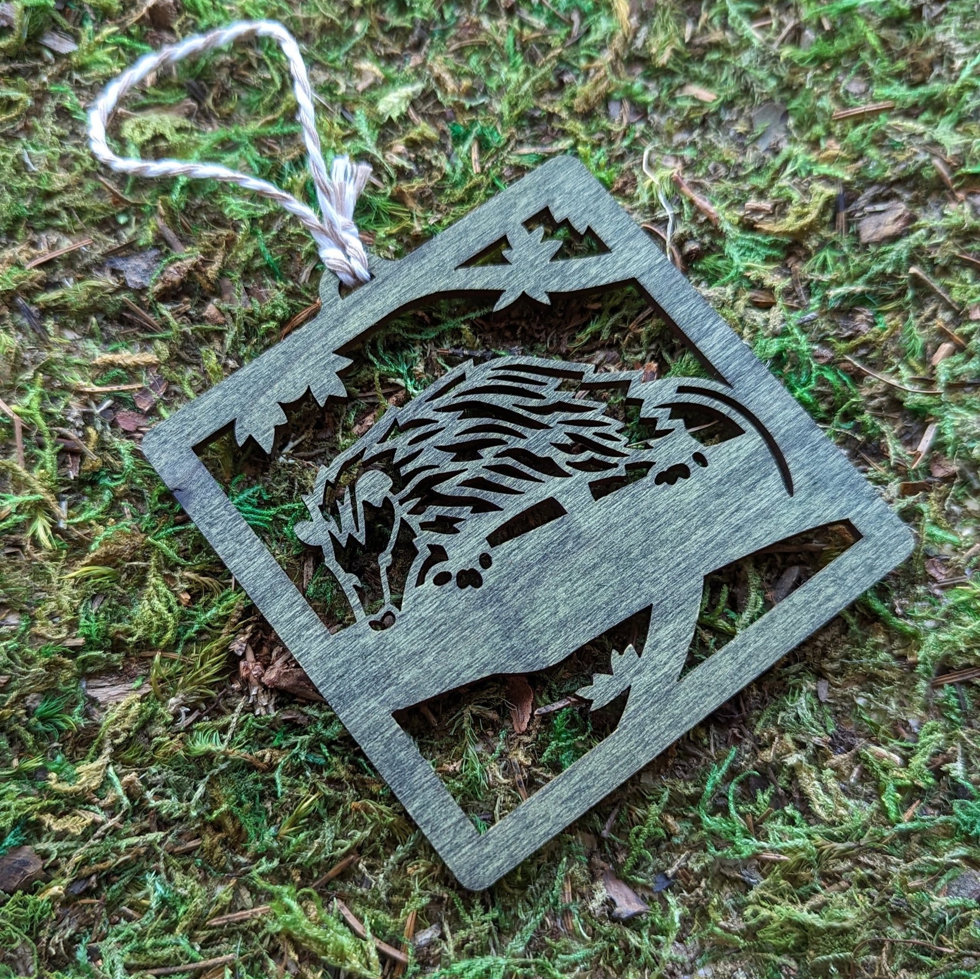 A wooden laser cut ornament of an opossum on a tree branch colored with green wood dye on a mossy background.