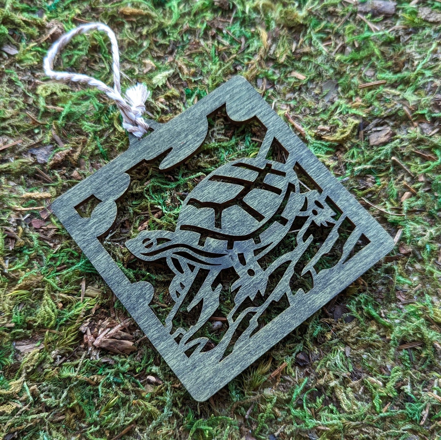 A wooden laser cut ornament of a turtle sitting on a log colored with green wood dye on a mossy background.