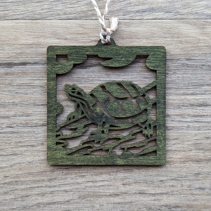 A wooden laser cut ornament of a turtle sitting on a log colored with green wood dye on a gray wood background.