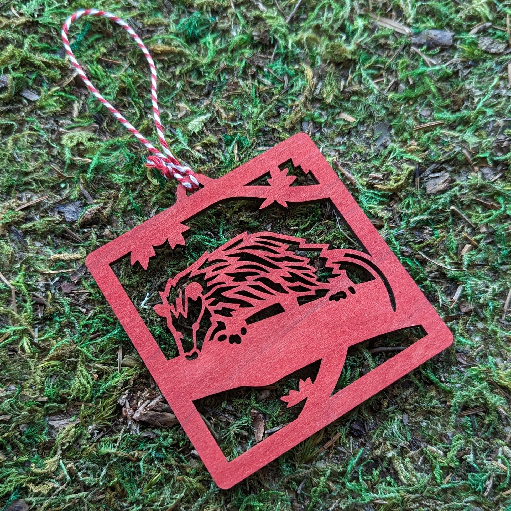 A wooden laser cut ornament of an opossum on a tree branch colored with red wood dye on a mossy background.