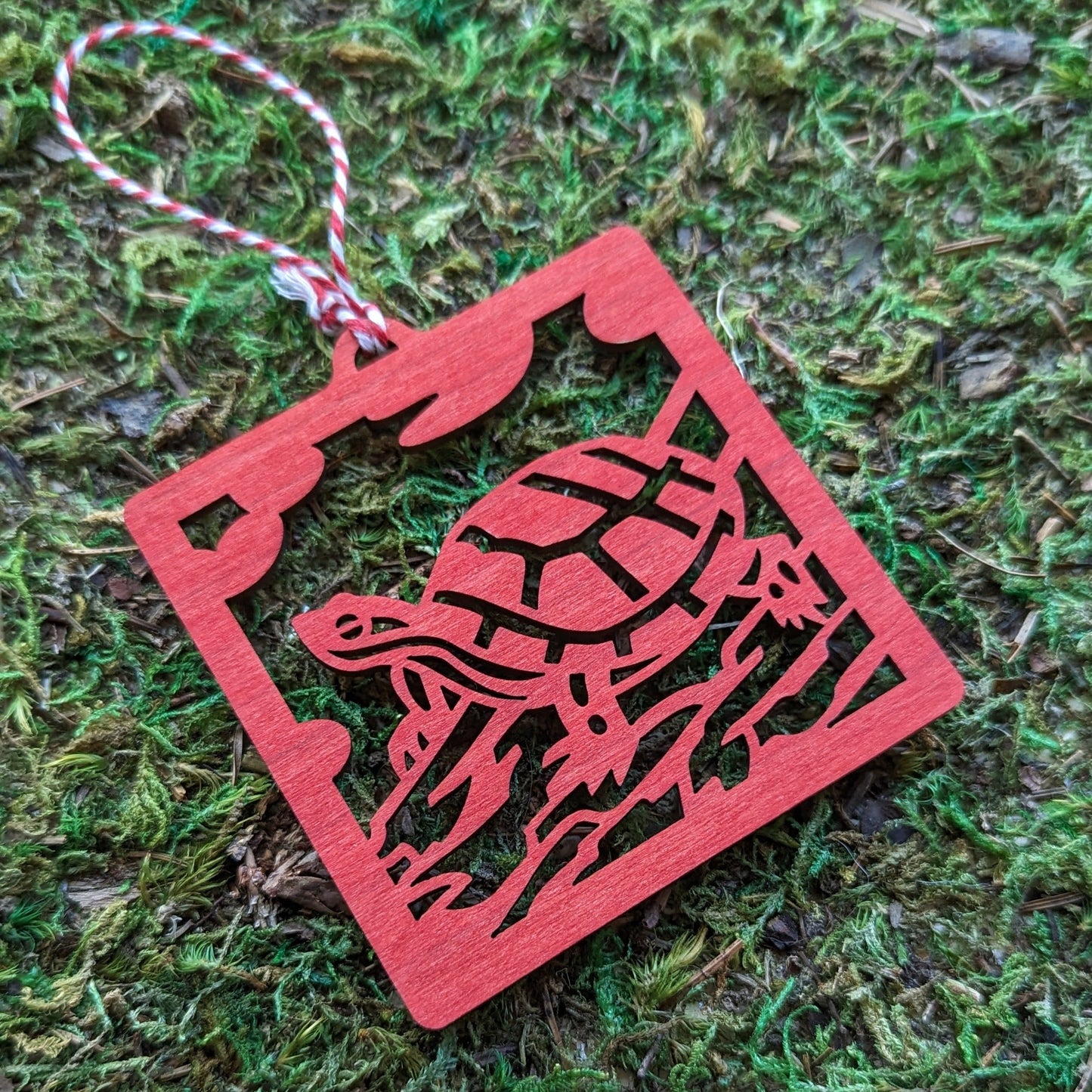 A wooden laser cut ornament of a turtle sitting on a log colored with red wood dye on a mossy background.