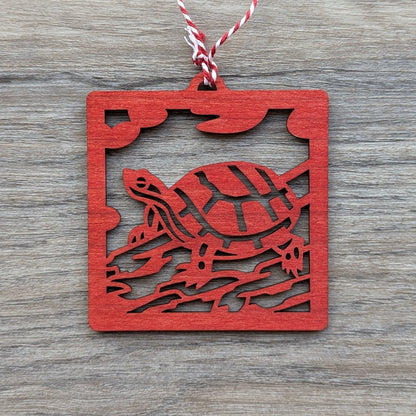 A wooden laser cut ornament of a turtle sitting on a log colored with red wood dye on a gray wood background.