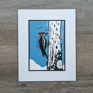 An original screen print of an illustration of a pileated woodpecker perched on a tree and pecking at the bark.